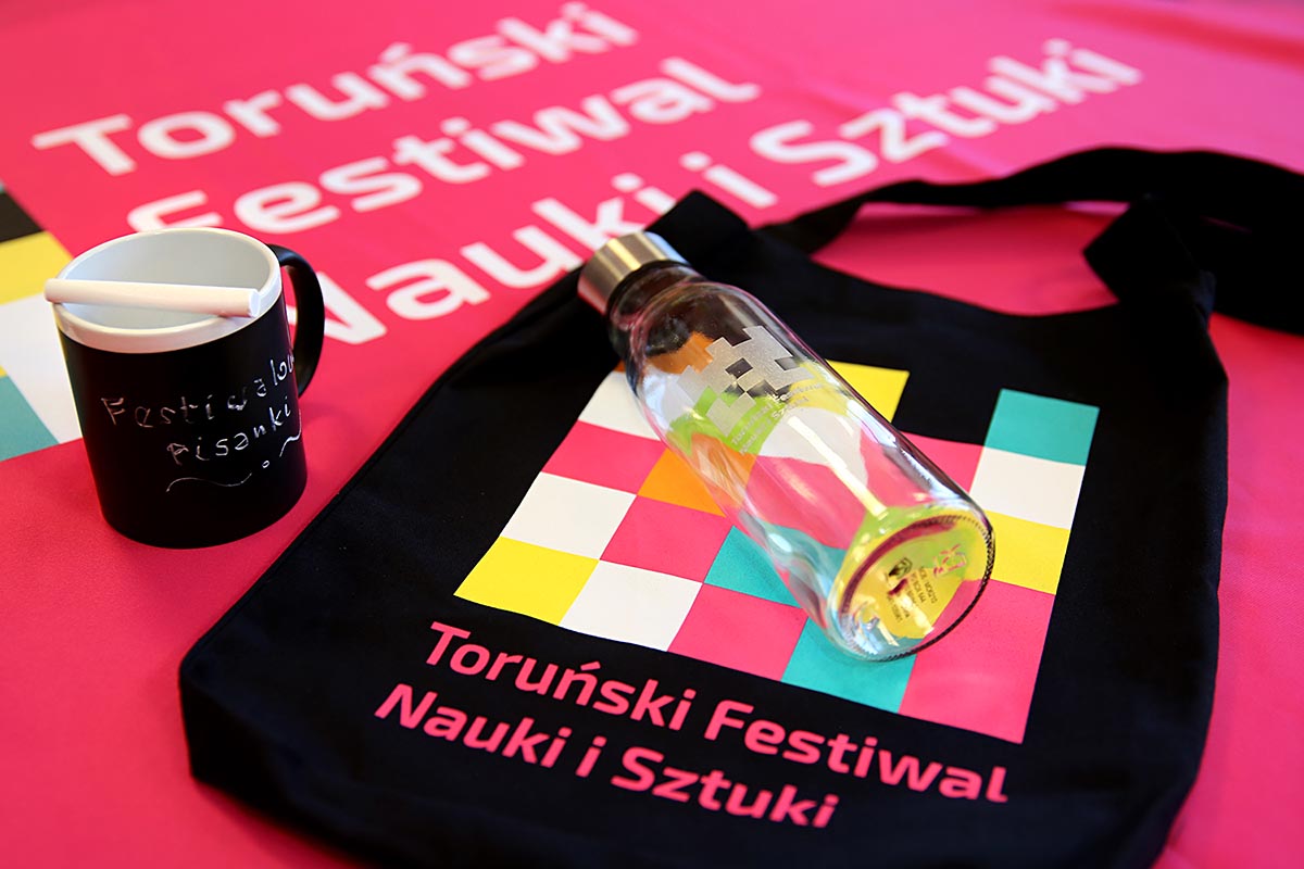 Zdjęcie przedstawiające nagordy konkursowe: czarna torba z logo Festiwalu, czarny kubek, szklana butelka.